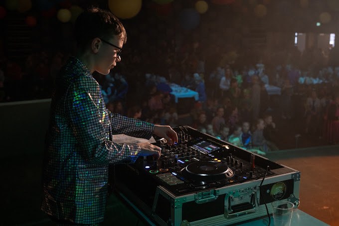 DJ Nori in een glimmend jasje achter de draaitafel, met een volle zaal op de achtergrond